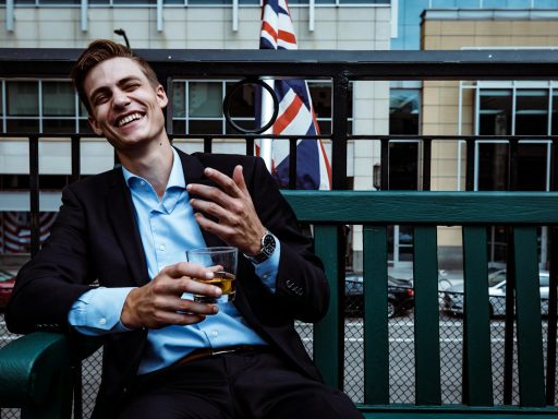 man smiling while sitting and holding whisky glass near concrete building